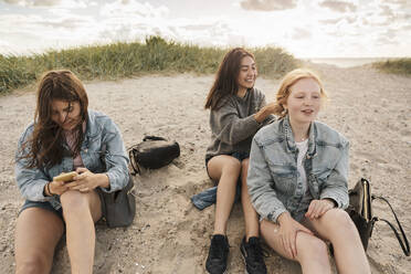 Teenage girl using smart phone while female friends sitting on sand at beach - MASF23509