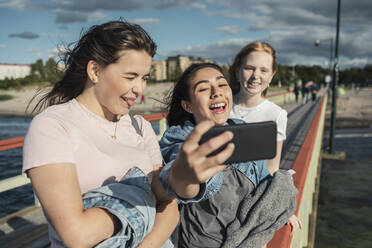 Fröhliche Freundinnen nehmen Selfie durch Smartphone auf Pier am Meer während sonnigen Tag - MASF23510