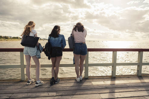Rückansicht von Freundinnen, die sich bei Sonnenuntergang am Geländer des Piers anlehnen - MASF23513