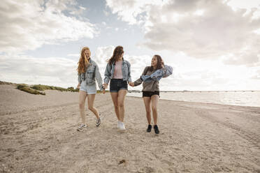 Happy female friends holding hands while walking on sand at beach - MASF23515