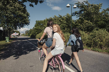 Teenage girl walking by female friends cycling on road during sunny day - MASF23520