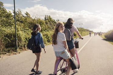 Female friends cycling while teenage girl walking on road during sunny day against sky - MASF23523