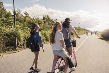 Weibliche Freunde auf dem Fahrrad, während ein Teenager-Mädchen auf der Straße an einem sonnigen Tag gegen den Himmel läuft - MASF23523