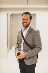 Portrait of smiling male entrepreneur with smart phone in corridor at office - MASF23531