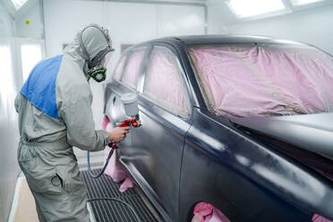 Side view of male in respiratory mask and protective suit painting car with spray gun in service - ADSF24170