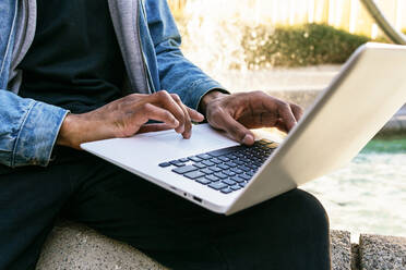 Cropped unerkennbar ethnischen afroamerikanischen erwachsenen männlichen Remote-Mitarbeiter mit Laptop in der Stadt sitzen - ADSF24171