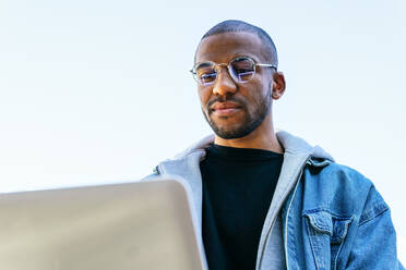 From below ethnic African american adult male remote employee with laptop sitting in city - ADSF24172