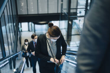 Female entrepreneur peeking in sling bag while standing on escalator during pandemic - MASF23559