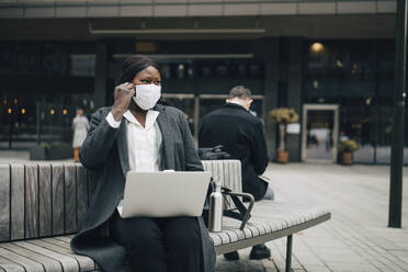 Businesswoman with laptop looking away while sitting on bench during pandemic - MASF23566