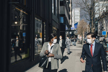 Male and female colleagues discussing while walking on sunny day in city during COVID-19 - MASF23602