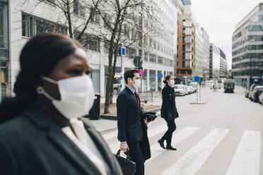 Female and male colleagues crossing street in city during pandemic - MASF23612