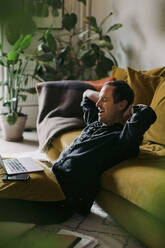 Male entrepreneur yawning with hands behind head in living room at home - MASF23674