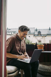 Lächelnder Geschäftsmann, der auf dem Balkon sitzend am Laptop arbeitet, während COVID-19 - MASF23678