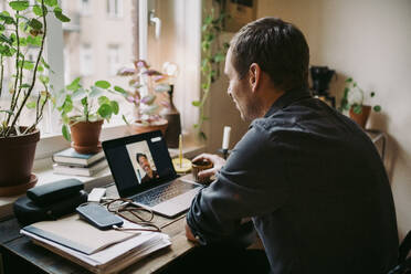 Businessman with coffee cup discussing with colleague on video call at home office - MASF23707