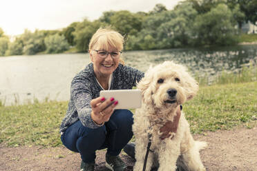 Lächelnde Frau nimmt Selfie mit Hund durch Smartphone am Seeufer - MASF23776