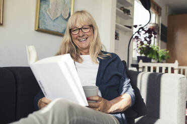 Lächelnde Frau liest ein Buch, während sie zu Hause auf dem Sofa sitzt - MASF23788