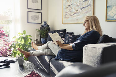 Blond woman reading book while sitting on sofa in living room - MASF23789