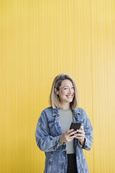 Lächelnde Frau mit Mobiltelefon vor einer Wand stehend - EBBF03440