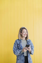 Smiling woman with mobile phone standing in front of wall - EBBF03440