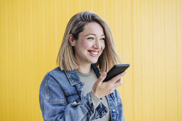 Glückliche Frau, die eine Sprachnachricht über ihr Smartphone an eine gelbe Wand sendet - EBBF03441