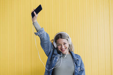 Happy woman with mobile phone listening music through headphones in front of yellow wall - EBBF03446