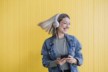 Happy woman with tousled hair looking away while holding mobile phone in front of wall - EBBF03447