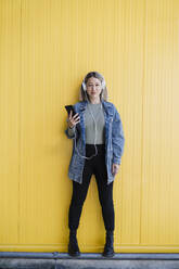 Woman wearing headphones standing at retaining wall - EBBF03449