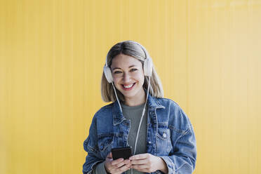 Glückliche blonde Frau hält ihr Smartphone in der Hand und hört Musik über Kopfhörer vor einer gelben Wand - EBBF03450
