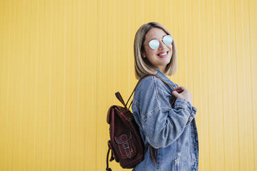 Lächelnde blonde Frau mit Rucksack und Sonnenbrille an einer gelben Wand - EBBF03451