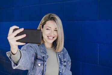 Frau nimmt Selfie durch Smartphone durch blaue Wand - EBBF03453
