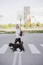 Lächelnde Frau mit Hund beim Überqueren der Straße in der Stadt - EBBF03465