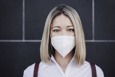 Blonde Frau mit Gesichtsschutzmaske vor einer Wand während einer Pandemie - EBBF03466