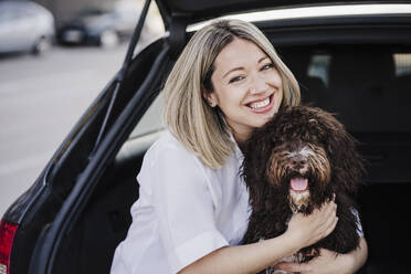 Glückliche Frau umarmt spanischen Wasserhund im Kofferraum eines Autos - EBBF03467
