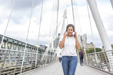 Lächelnde junge Frau, die beim Gehen auf einer Brücke über Kopfhörer Musik hört - WPEF04436