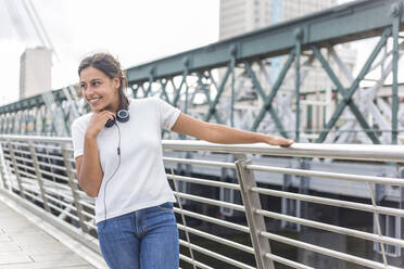 Lächelnde junge Frau, die wegschaut, während sie am Geländer einer Brücke steht - WPEF04439