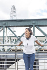 Glückliche Frau mit Kopfhörern, die vor einem Brückengeländer steht, London, England, UK - WPEF04441