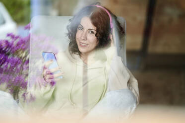 Lächelnde Frau mit Mobiltelefon durch ein Fenster gesehen - KIJF03831