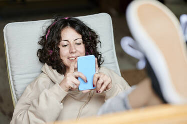 Lächelnde Frau mit hochgelegten Füßen, die zu Hause ein Mobiltelefon benutzt - KIJF03832