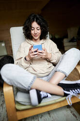 Lächelnde Frau sitzt mit gekreuzten Beinen und benutzt ein Mobiltelefon zu Hause - KIJF03838