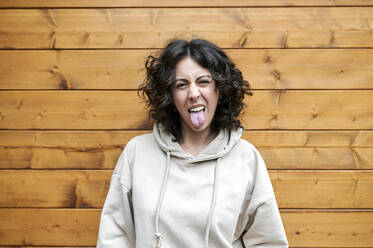 Lustige Frau, die ihre Zunge herausstreckt, während sie vor einer Holzwand steht - KIJF03851