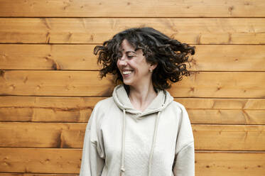 Lächelnde Frau, die sich die Haare zuwirft, während sie vor einer Holzwand steht - KIJF03852