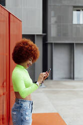 Happy woman using mobile phone in front of red wall - PGF00558