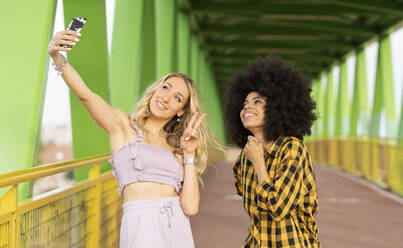 Female friends taking selfie through smart phone on bridge - JCCMF02329