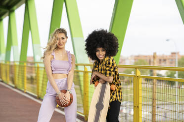 Multi-ethnische Freundinnen mit Skateboard und Basketball auf einer Brücke stehend - JCCMF02333