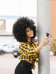 Afro-Frau, die ein Selfie mit ihrem Smartphone macht, während sie sich an einen Pfahl lehnt - JCCMF02349