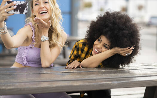 Lachende Freundinnen während eines Videoanrufs mit dem Smartphone am Tisch - JCCMF02363
