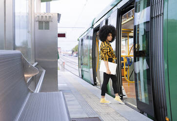 Afro woman holding laptop while entering in train at railroad station - JCCMF02382