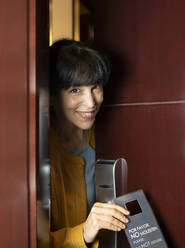 Lächelnde Frau späht durch die Tür einer Hotelsuite - JCCMF02391