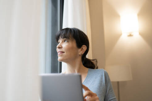 Ältere Frau, die durch das Fenster schaut, während sie ein digitales Tablet im Hotelzimmer benutzt - JCCMF02396