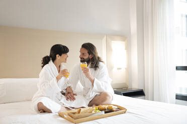 Paar in Bademänteln trinkt Saft auf dem Bett einer Hotelsuite - JCCMF02414